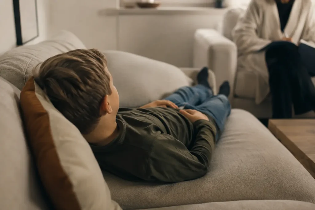 Dreng ligger afslappet på briks med lukkede øjne under klinisk hypnosebehandling, mens behandler sidder i baggrunden